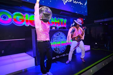 Boogie Nights with Disco Ball on the dance stage with arm raised dancing along side a person in costume.