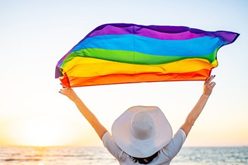 Woman standing with back turned in front of beach holding a translucent pride flag and wearing a white brimmed hat.