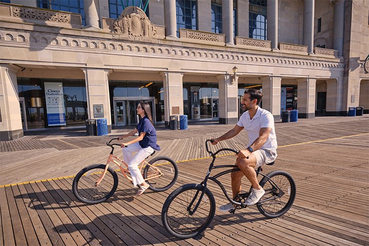 Man and woman bike riding on the Atlantic City Boardwalk with Jim Whelan Boardwalk Hall in background.